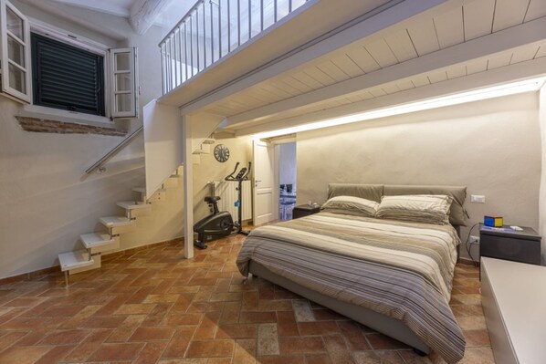 A bedroom with a bed, a nightstand, a desk, a window, and a staircase leading to an upper floor.