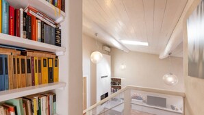 Een modern interieur met witte wanden, een houten plafond en een trap. Links bevinden zich boekenkasten en in het midden een eethoek met een tafel en stoelen.