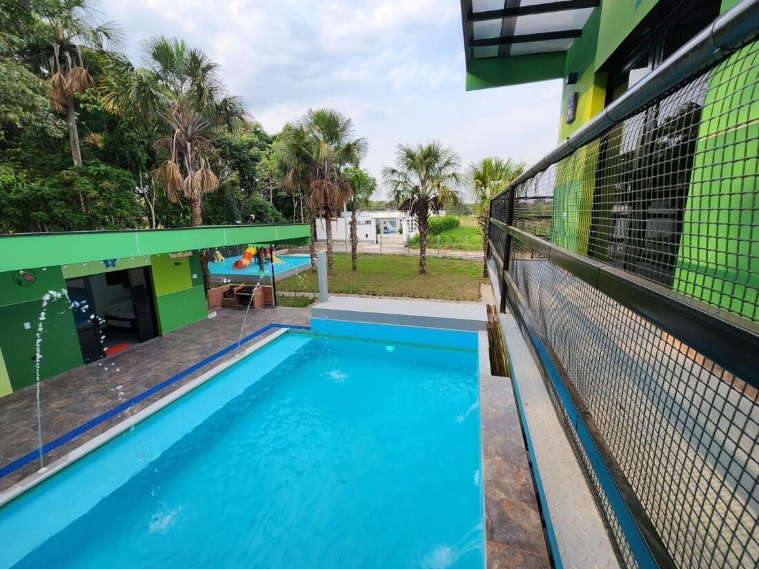 A swimming pool with a clear blue water, surrounded by a green fence, and palm trees in the background.