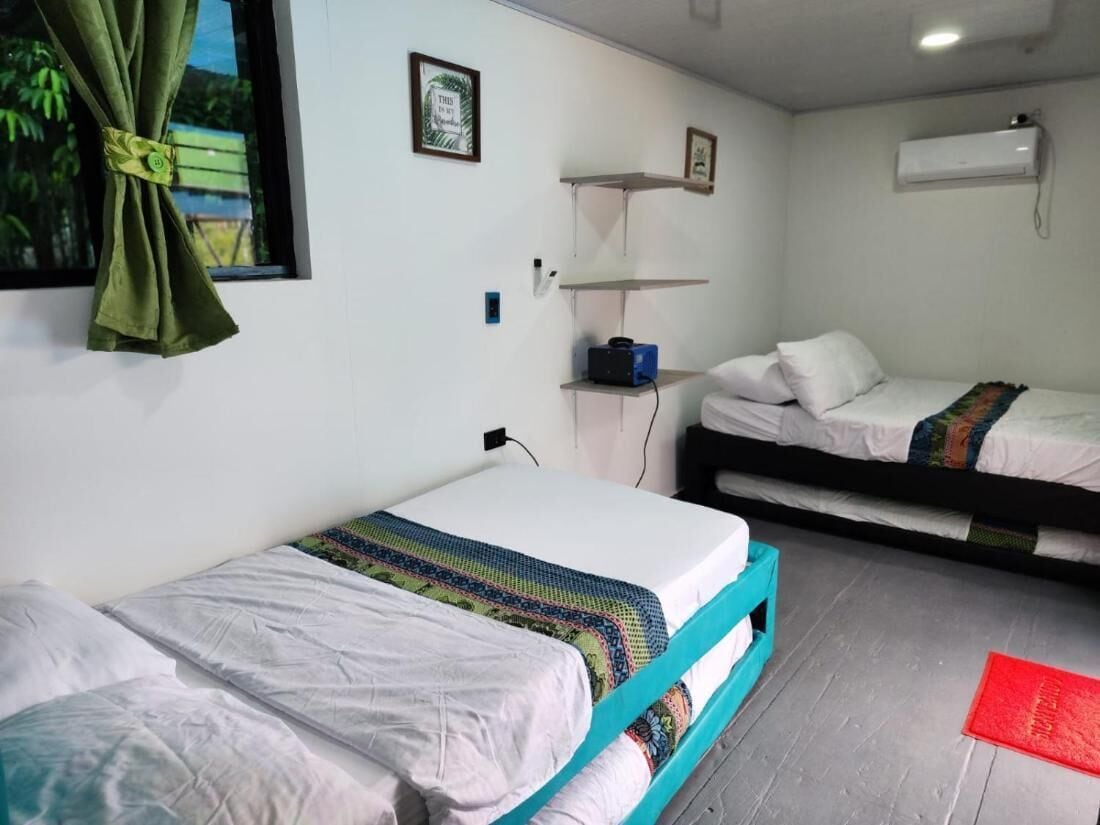 A room with two beds, a window with curtains, a shelf with a small item, and an air conditioning unit.
