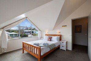 A bedroom with a sloped ceiling, a large window, a bed with pillows, bedside tables, and a door. - A Hop  Skip & Jump to the Lake (Taupo)