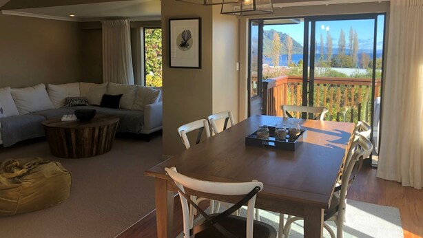 A spacious living area with a wooden dining table, white chairs, a sofa, and a coffee table. There is a large window with a view of a deck and mountains.