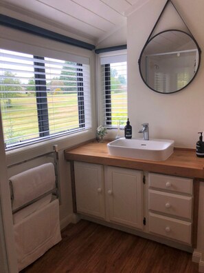 A bathroom with a white sink, wooden countertop, and a mirror. There are two windows with blinds, and a towel rack is visible.