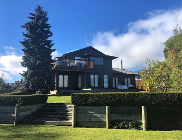 A modern two-story house with a balcony, surrounded by well-maintained hedges and a large evergreen tree.