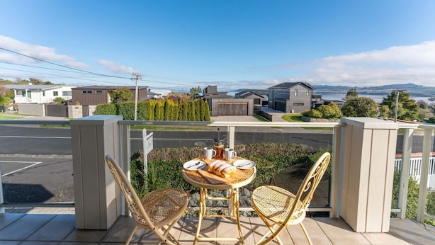 A balcony with a table and chairs, overlooking a residential area with houses and a body of water. - Top of the Lake in Taupo (Taupo)
