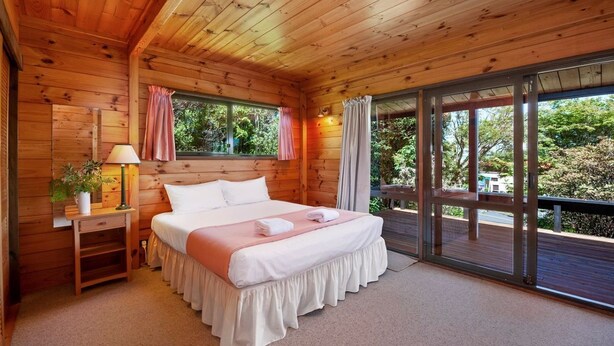 Un dormitorio de cabaña de madera con una cama, mesita de noche, lámpara y ventana con vistas a la vegetación.