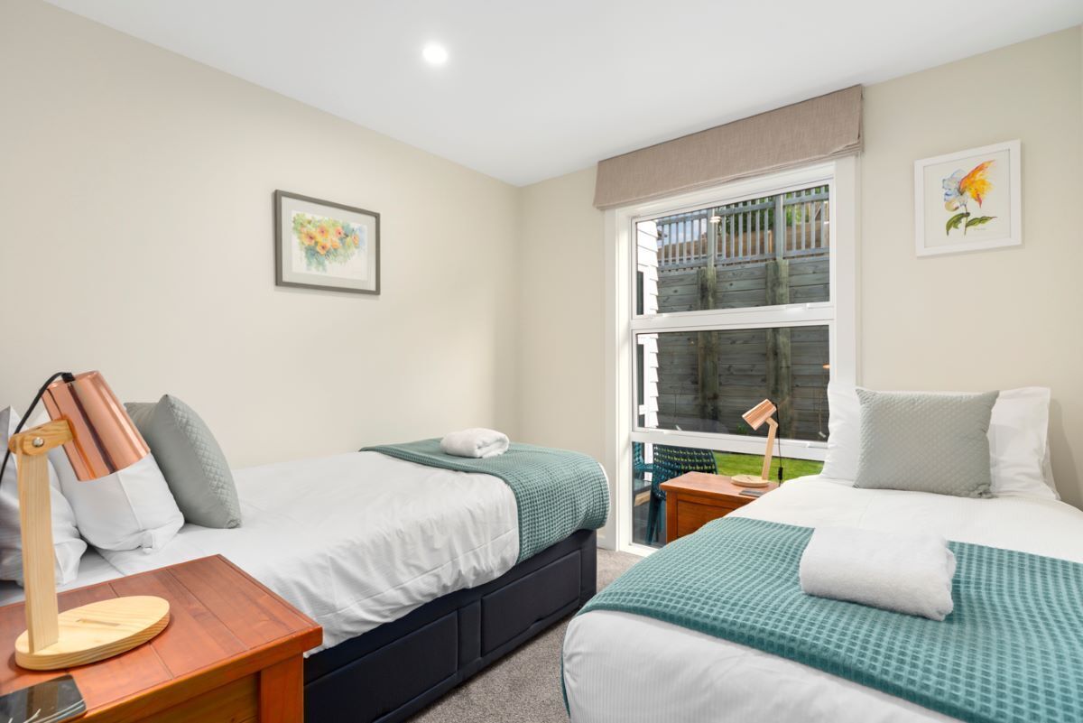 Quarto de hotel com duas camas, uma escrivaninha com abajur, janela com vista para uma varanda e um quadro na parede.