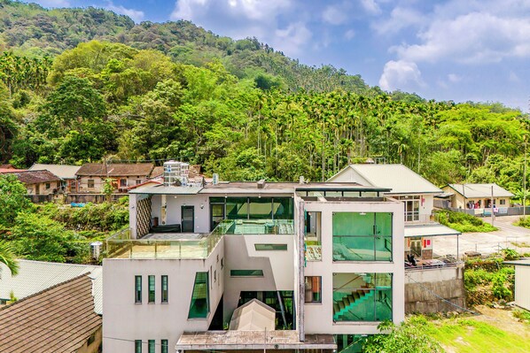 Un edificio moderno de varios pisos con fachada de vidrio, rodeado de vegetación exuberante y colinas al fondo.