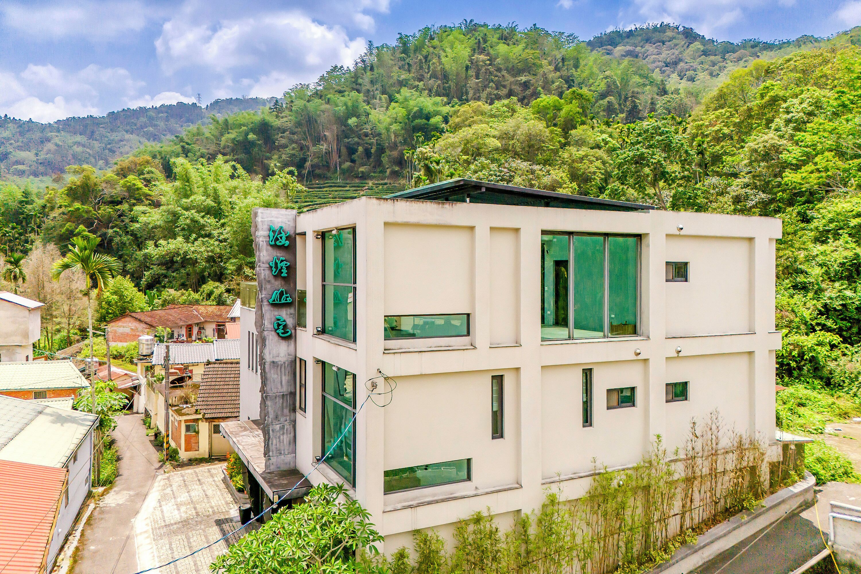 Un edificio moderno de dos plantas, con numerosas ventanas y techo plano, rodeado de abundante vegetación y colinas al fondo.