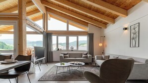 A spacious living area with a high ceiling, large windows offering a view of the outdoors, and a modern interior design.