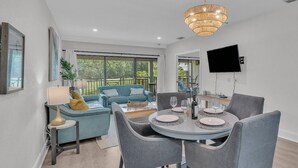 A modern living room with a dining area, large windows, and a flat-screen TV mounted on the wall.
