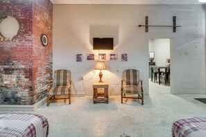 Una habitación con dos sillones de cuadros, una pequeña mesa de madera con una lámpara, un televisor montado en la pared y una pared de ladrillo con un ventanal circular.