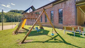 Ein Spielplatz mit Schaukeln und Rutsche, im Hintergrund ein Backsteinhaus und eine Rasenfläche.