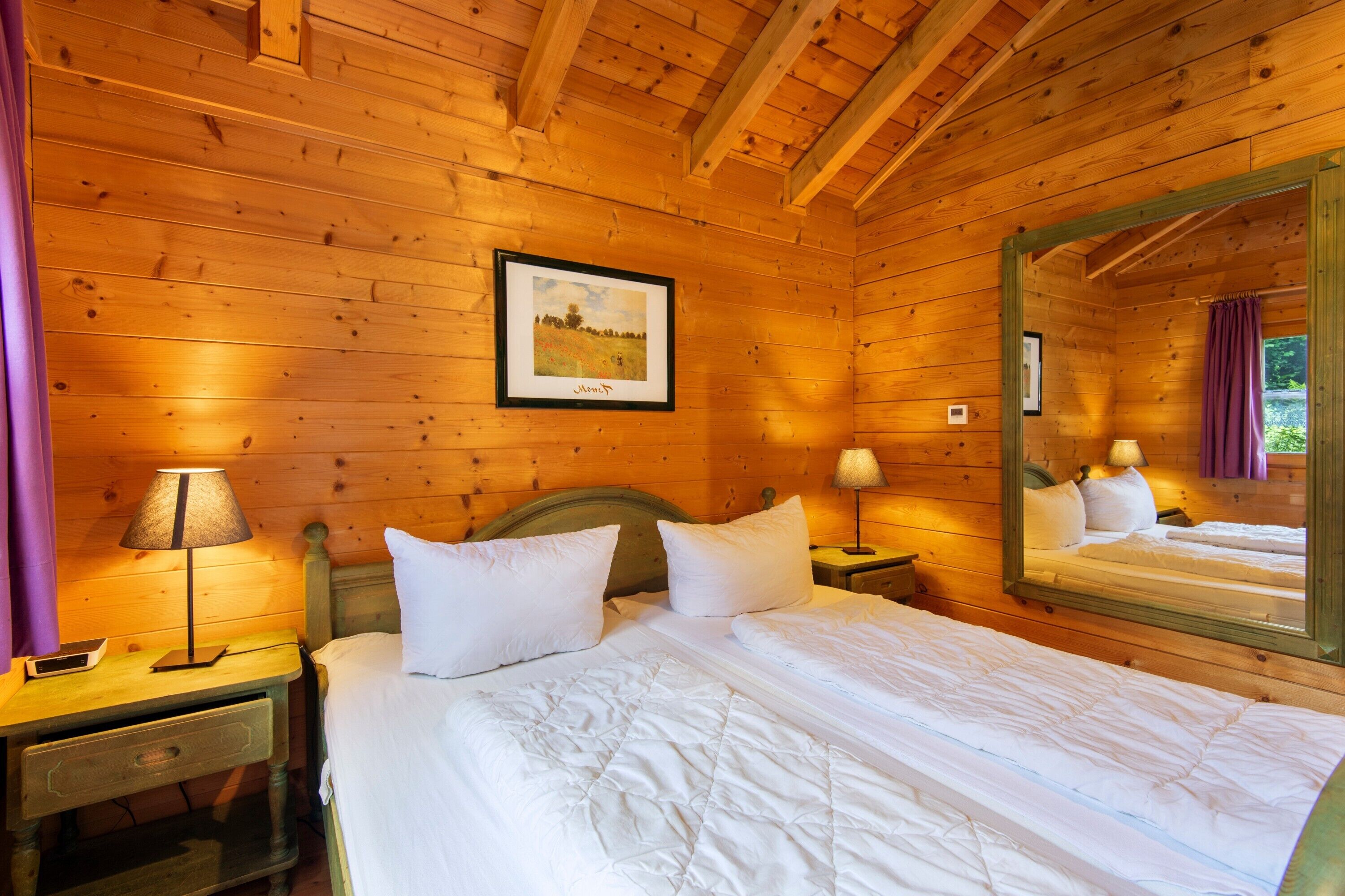 A wooden cabin room with a bed, two bedside tables, a lamp, a framed picture, and a mirror.