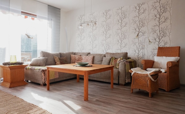 Una sala de estar con sofá, una mesa de centro de madera, una silla de mimbre y una pared decorada con motivos de árboles.