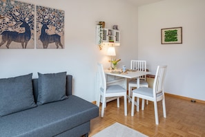 Ett vardagsrum med en grå soffa, ett matbord med vita stolar, ett litet skåp med växter och en inramad tavla på väggen.