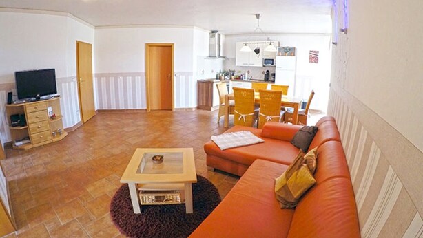 Una sala de estar con un sofá, una mesa de centro y un televisor. Se observa también una zona de cocina con una mesa y sillas para comer.