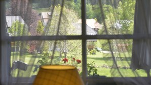 Una ventana con vista a un jardín y casas, una lámpara y un florero sobre una mesa.