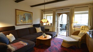 Un salón con sofá, sillón y una mesita de centro. Hay un cuadro en la pared y una ventana con cortinas.