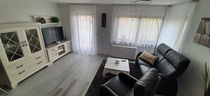 Un salon avec un canapé en cuir noir, une table basse blanche, un téléviseur posé sur un meuble et une fenêtre ornée de rideaux blancs.