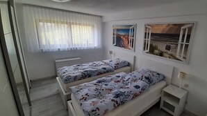 Une chambre avec deux lits, une fenêtre ornée de rideaux légers et deux tableaux accrochés au mur.