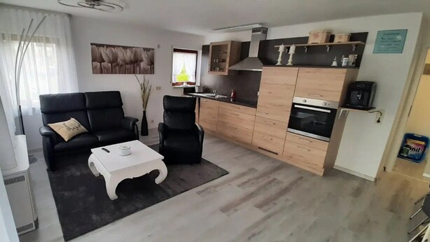 Un salon comprenant un canapé, des fauteuils, une table basse et un coin cuisine avec des placards et des appareils électroménagers.