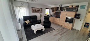 Een woonkamer met een bank, stoelen, een salontafel, en een keuken met kasten en apparatuur.