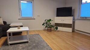 Ein Wohnzimmer mit einer Couch, einem kleinen Tisch, einer Topfpflanze, einem Schrank mit Fernseher und einem Fenster mit Jalousien.