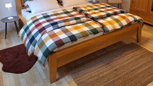 Ein Schlafzimmer mit einem hölzernen Bett, einer karierten Decke, einem Nachttisch mit Lampe, einem Sessel und einem Kleiderschrank.
