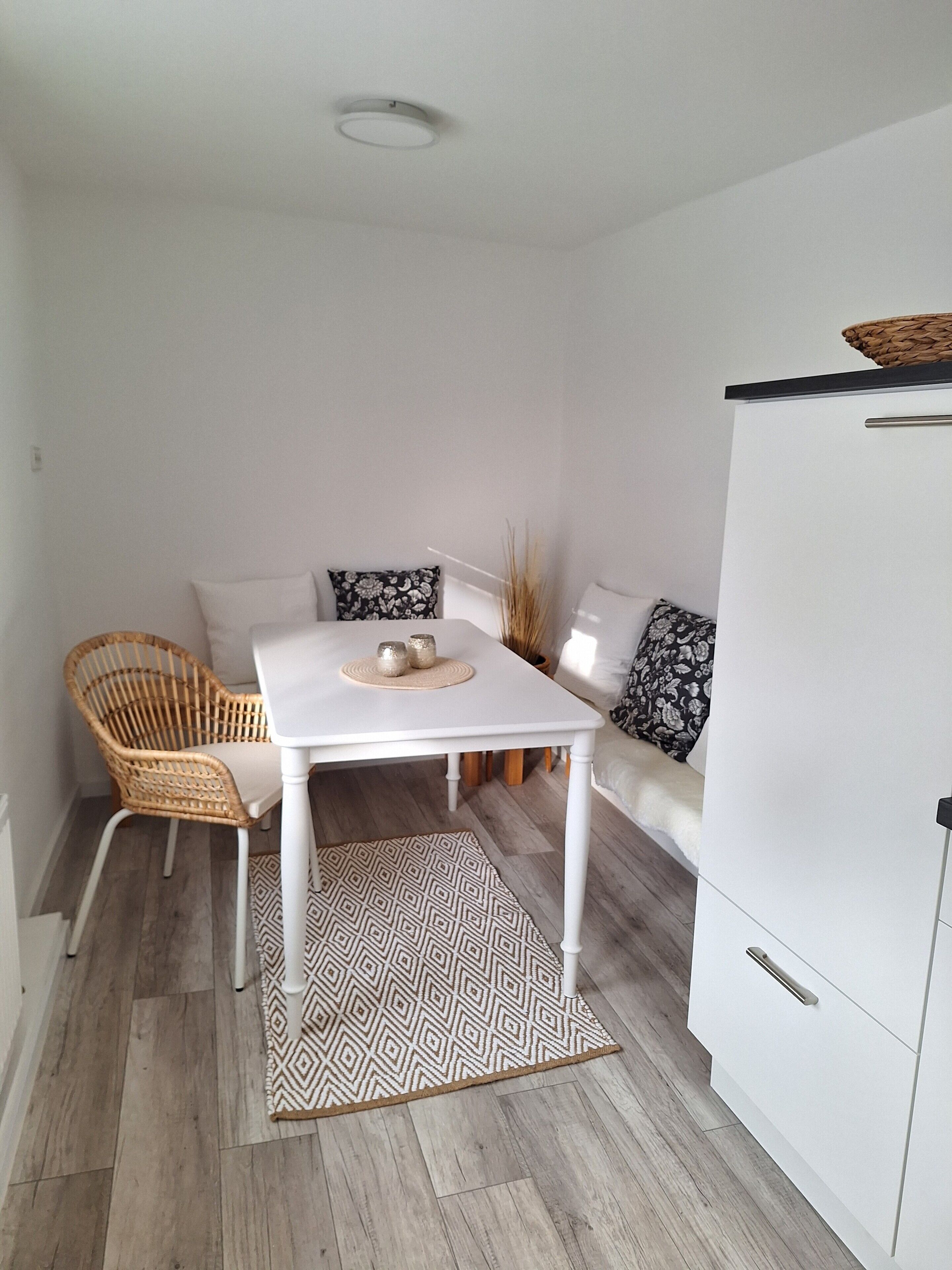 A room with a white table, wicker chair, patterned rug, and a sofa with decorative pillows.