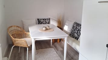A room with a white table, wicker chair, patterned rug, and a sofa with decorative pillows.