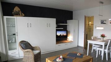 A modern living room with a television, wicker chair, dining table, and white cabinets.