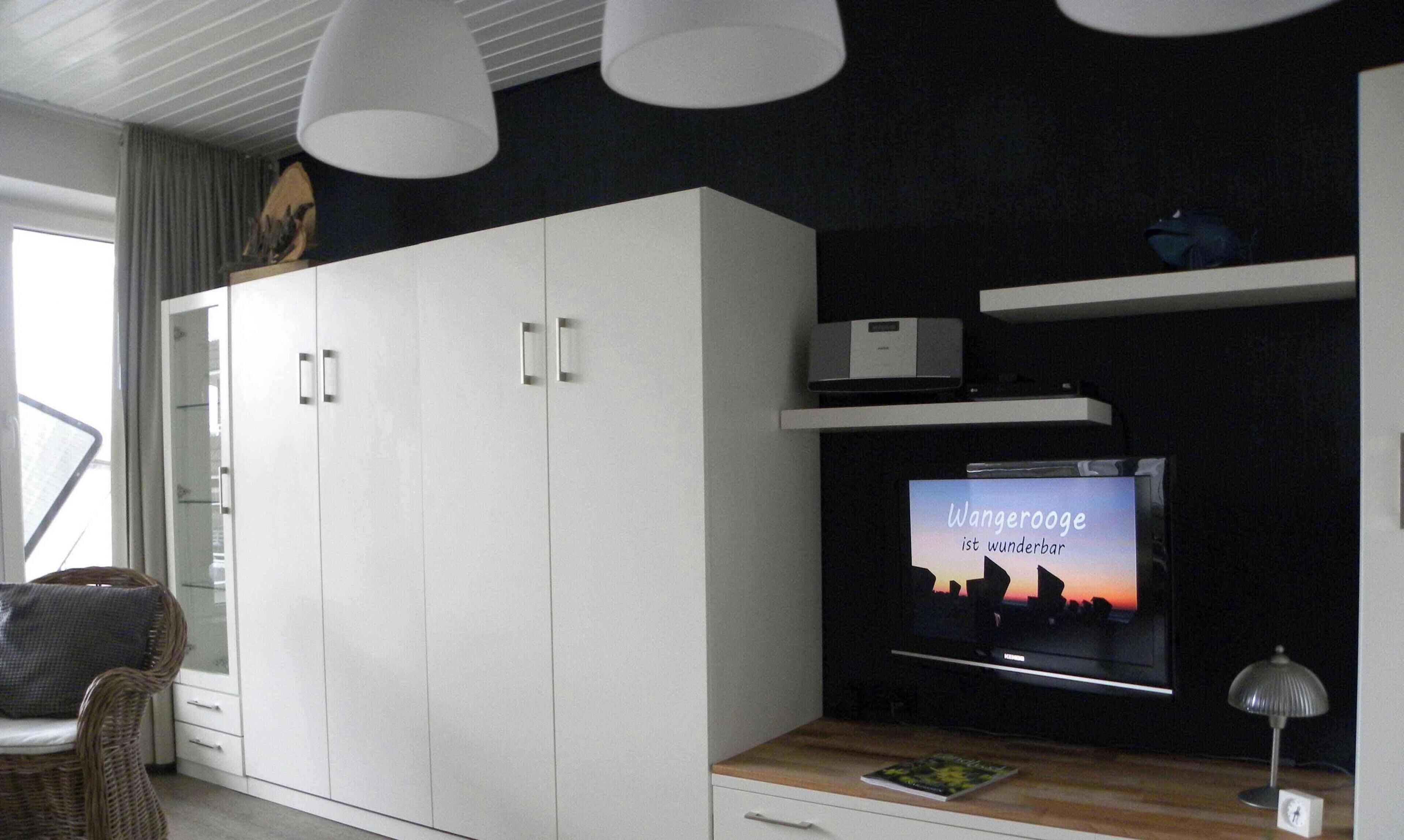 Una sala de estar moderna con televisor de pantalla plana, armarios blancos, una silla de mimbre e iluminación colgante.