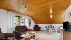 Un soggiorno con soffitto in legno, una zona pranzo con tavolo e sedie, un divano, una televisione e una finestra vista colline.