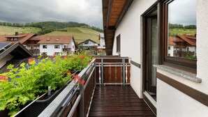 Un balcone con pavimento in legno, ringhiera in metallo e vista su case e vegetazione sullo sfondo.