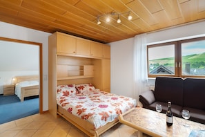 Quarto com cama, sofá, mesa com garrafa e copos, e janela com vista para colinas.