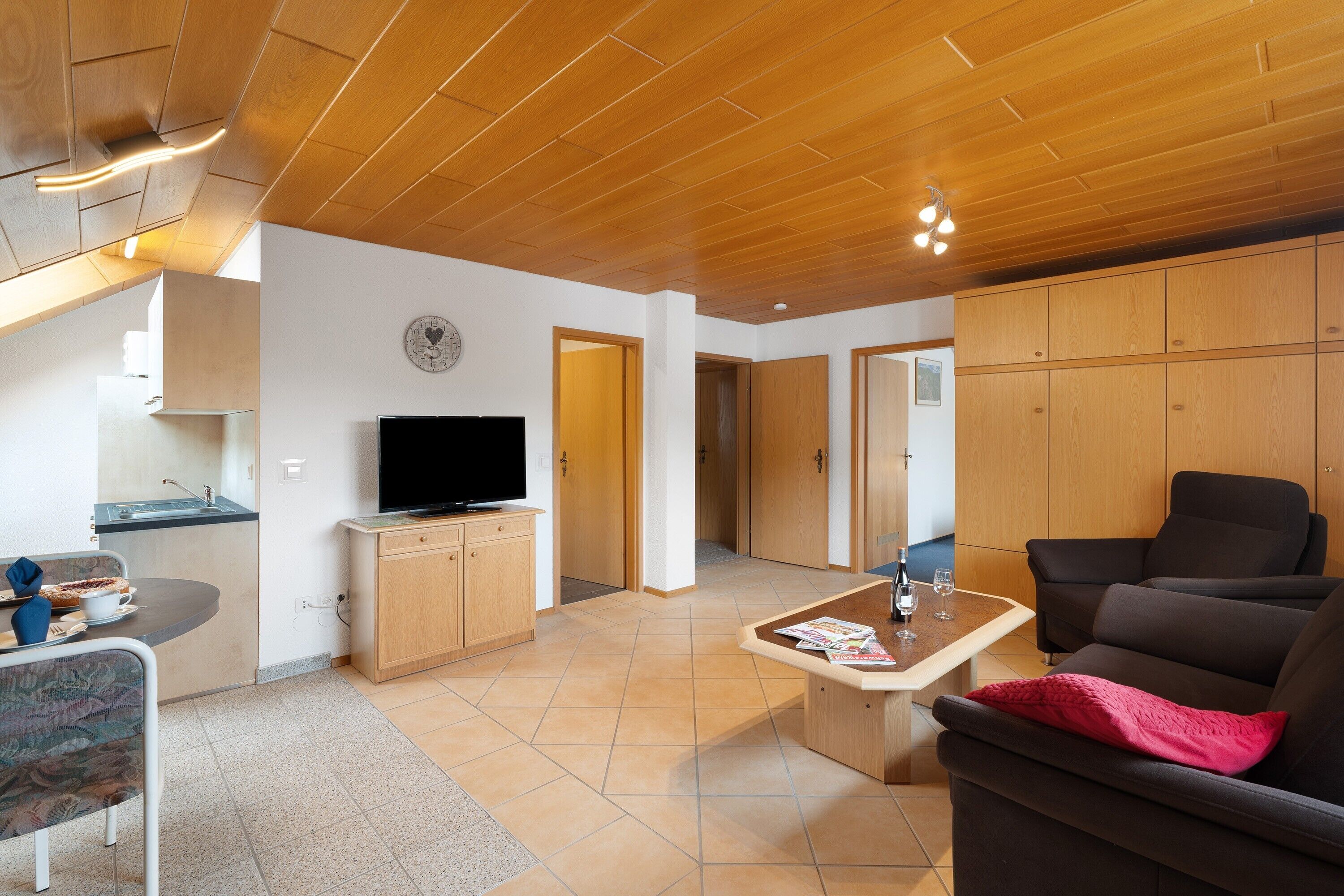 A modern living room with a flat-screen TV, wooden cabinets, a sofa, and a dining area with a table and chairs.