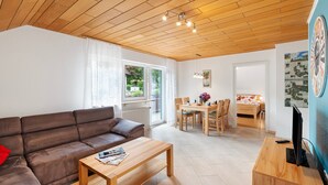 Ein modernes Wohnzimmer mit Holzboden, einem braunen Sofa, einem Holztisch, einer Essecke mit Tisch und Stühlen sowie einem Fernseher auf einem Holzschrank.
