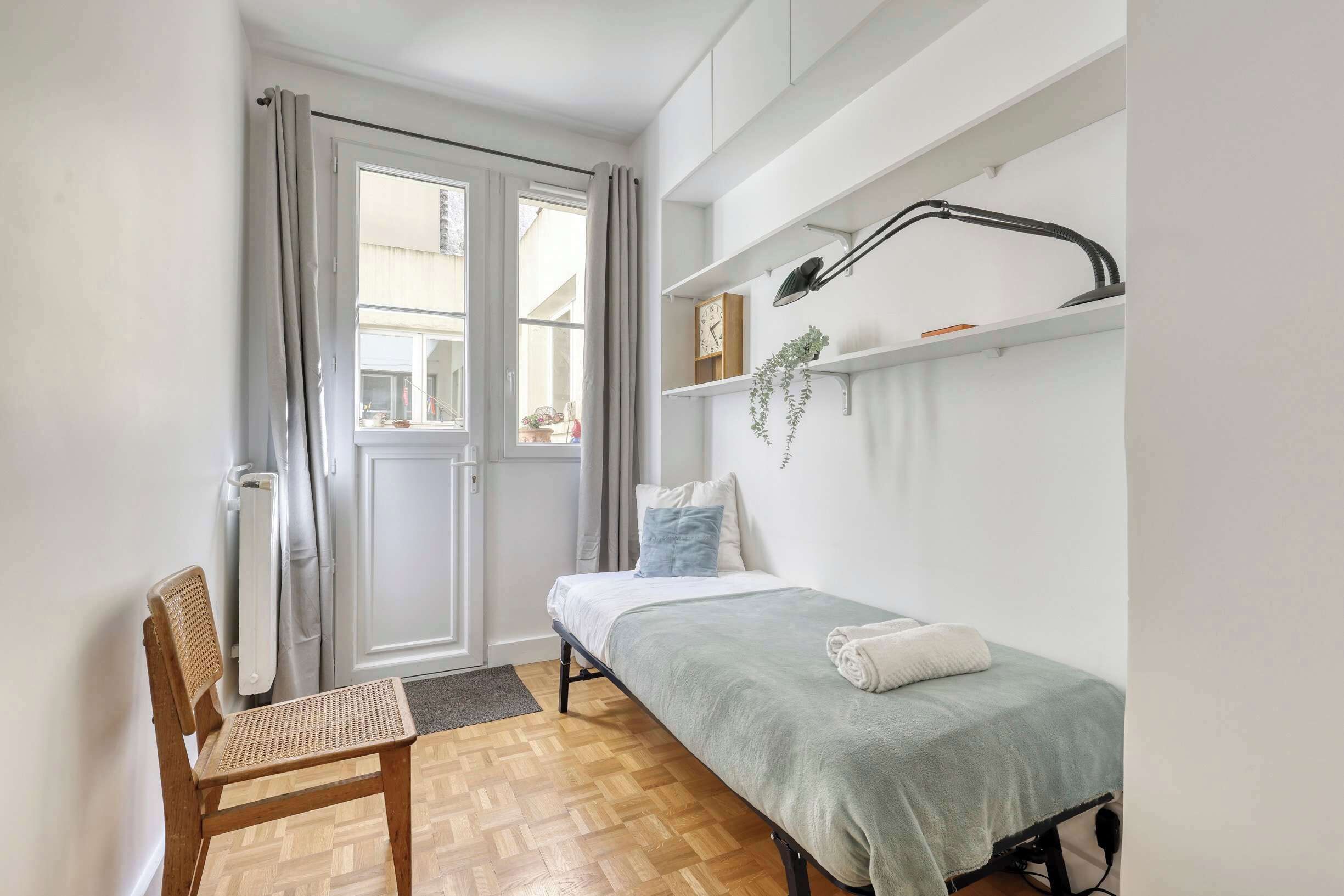 A small, well-lit room with a bed, a chair, a radiator, a window with curtains, and a shelf with a lamp and a clock.