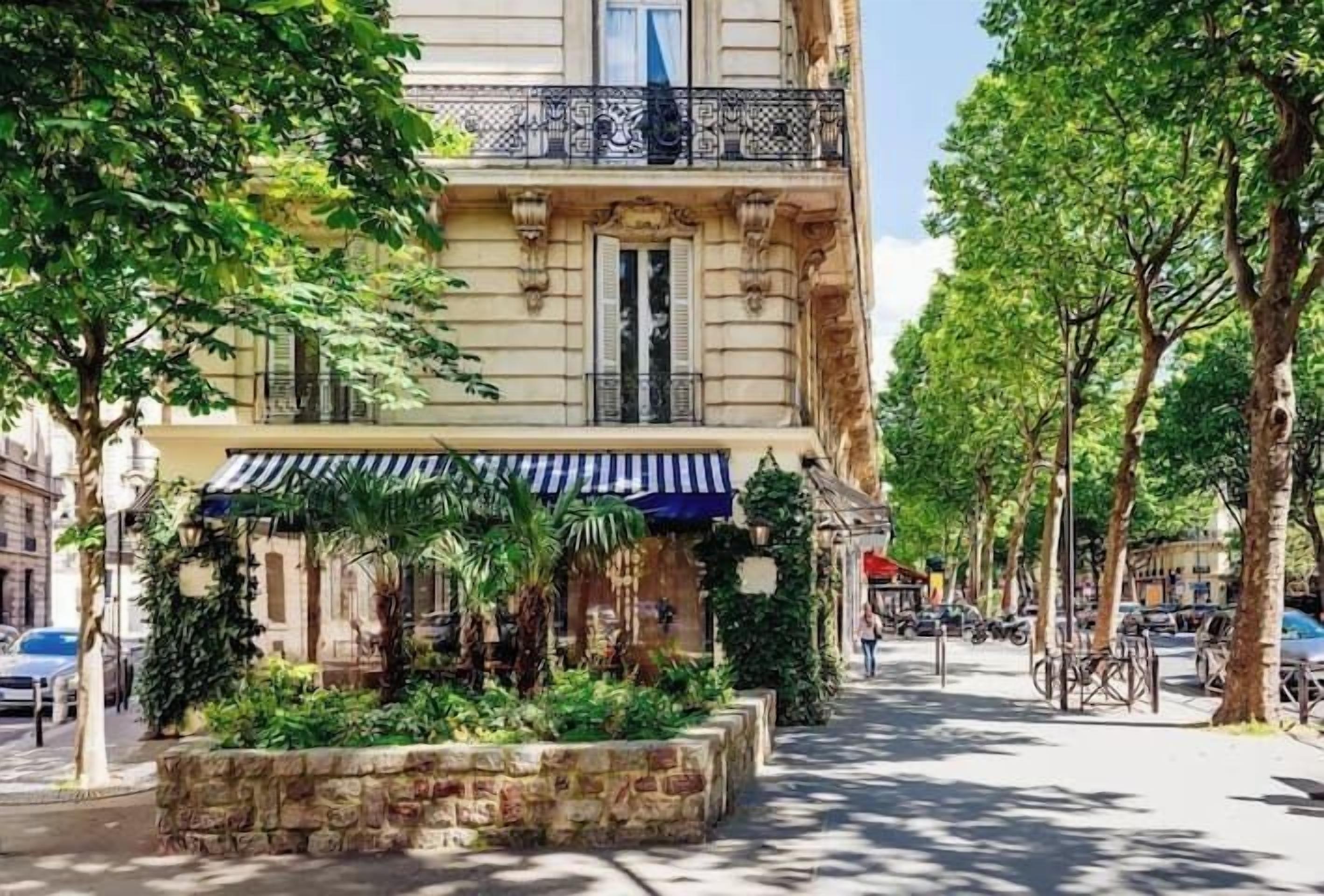 A historic building with a covered entrance, a stone planter with greenery, and a street with trees and parked cars.