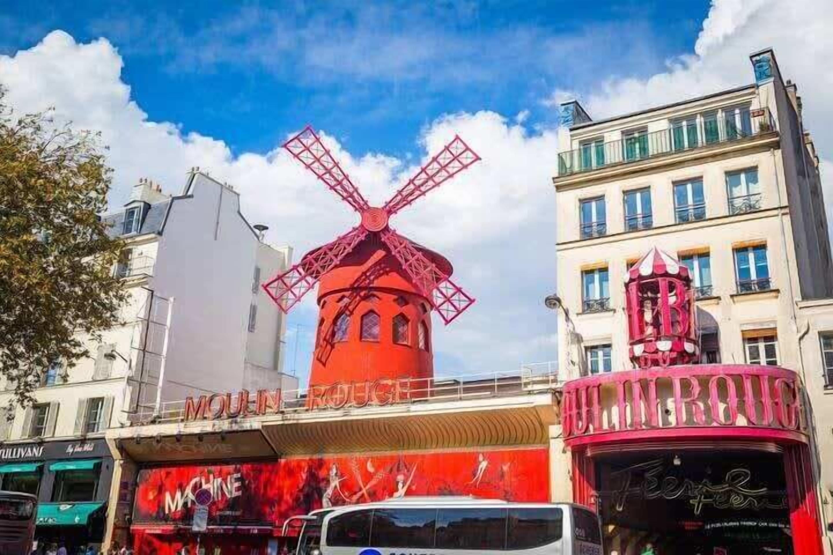 Un molino rojo con el letrero "Moulin Rouge" en la parte superior, rodeado de edificios y un autobús.