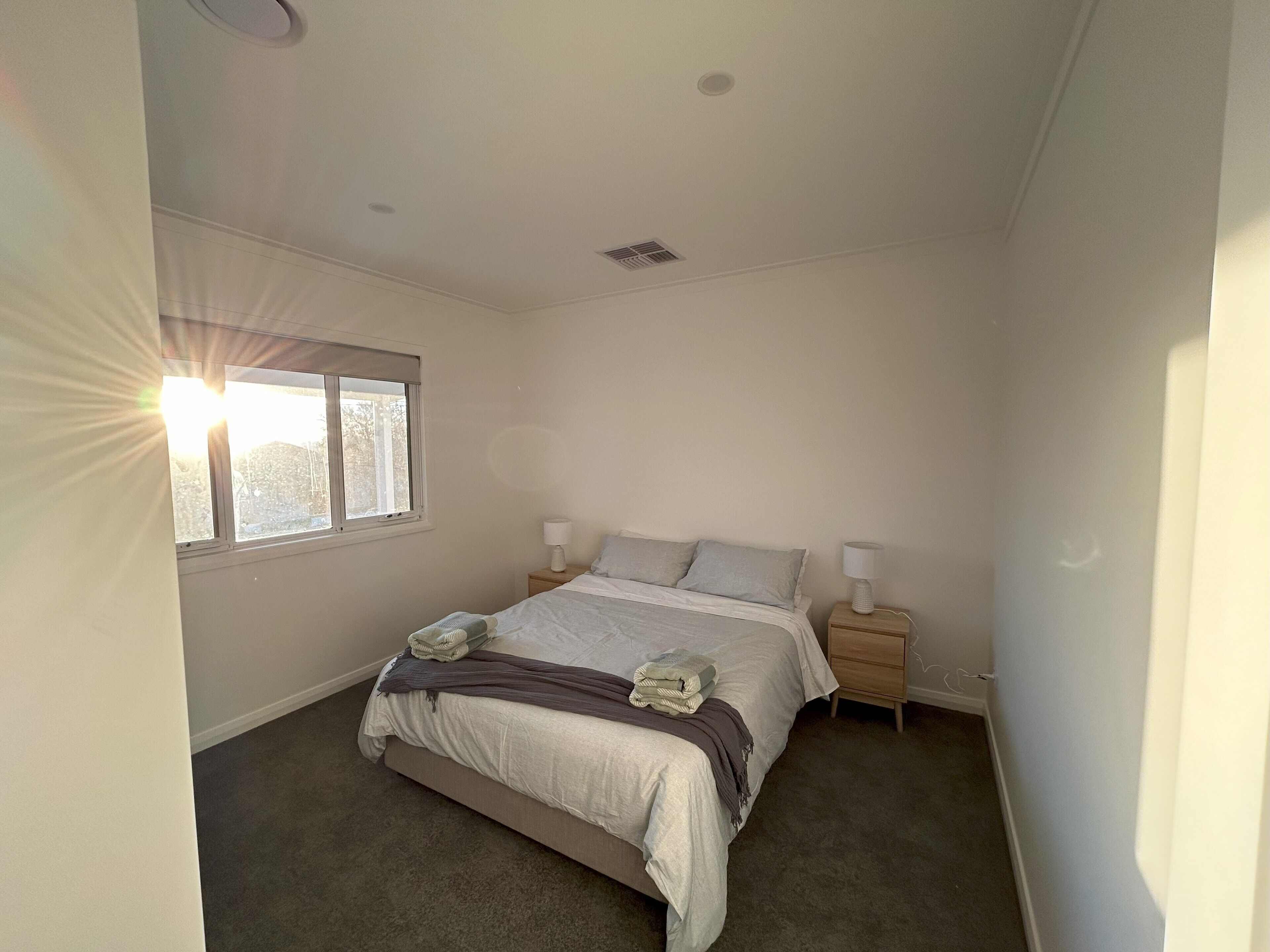 A bedroom with a bed, two bedside tables, a window, and a ceiling vent.