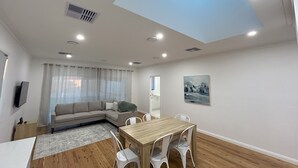 Un salon moderne avec une table à manger en bois, des chaises blanches, un canapé beige et une grande fenêtre ornée de rideaux légers.