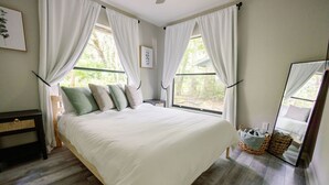 A bedroom with a large bed, white curtains, a bedside table, a nightstand, a mirror, and a window with a view of greenery. - Quiet 1-Story Townhome off I-75 by UF, Shands, VA, Celebration Pointe &Shopping! (Gainesville)