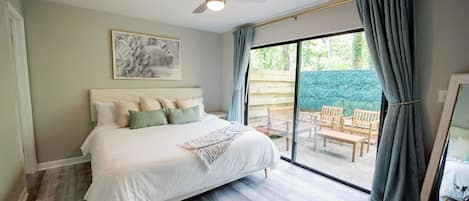 A bedroom with a bed, a ceiling fan, a large window with curtains, and a view of an outdoor patio area.
