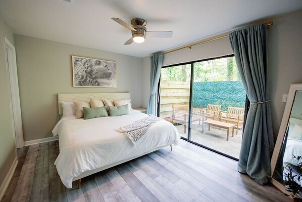 A bedroom with a bed, a ceiling fan, a large window with curtains, and a view of an outdoor patio area.