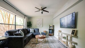 A modern living room with a sectional sofa, a round coffee table, a ceiling fan, a large TV, and a bookshelf. - Quiet 1-Story Townhome off I-75 by UF, Shands, VA, Celebration Pointe &Shopping! (Gainesville)