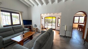 A modern living room with a grey sofa, a wooden coffee table, and a TV. There is a dining area with a wooden table and chairs, and a hallway leading to another room. - La Novela - Calpe, Costa Blanca (Calpe)