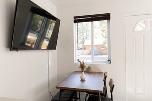 Una habitación con televisión montada en la pared, ventana con vistas al exterior, una mesa con sillas y una puerta.