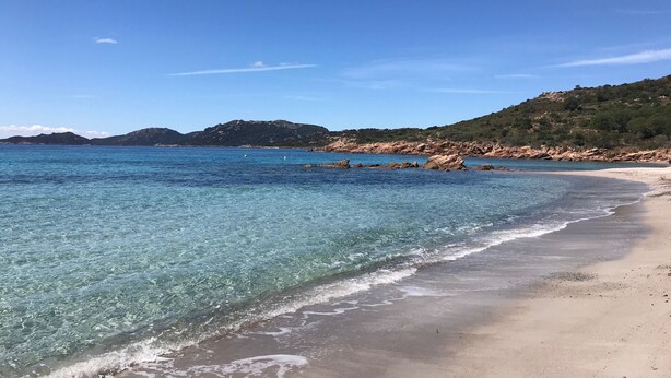 En strand med krystalklart blåt vand, sandet bredde og bakker i baggrunden.
